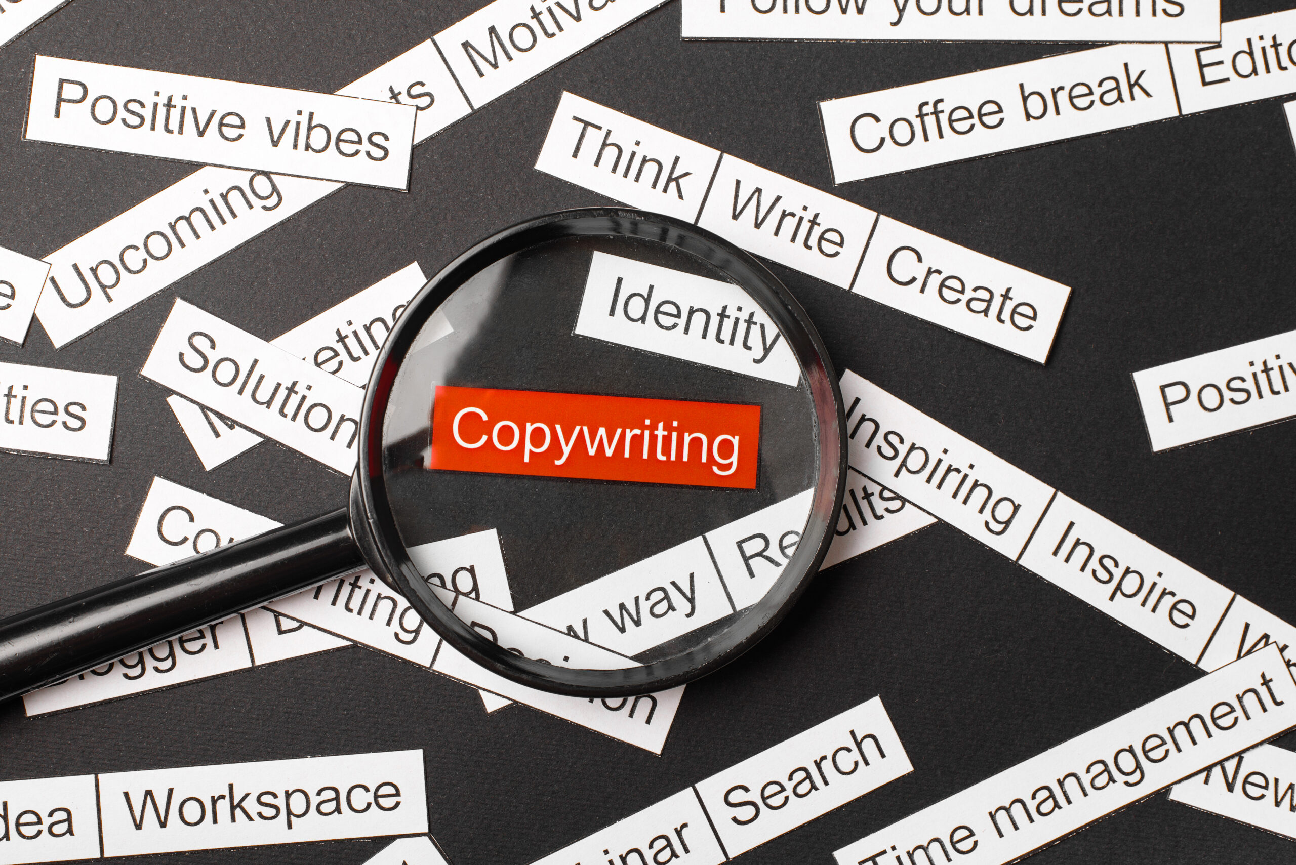 Magnifier glass over the red inscription copywriting cut out of paper. Surrounded by other inscriptions on a dark background. Word cloud concept.
