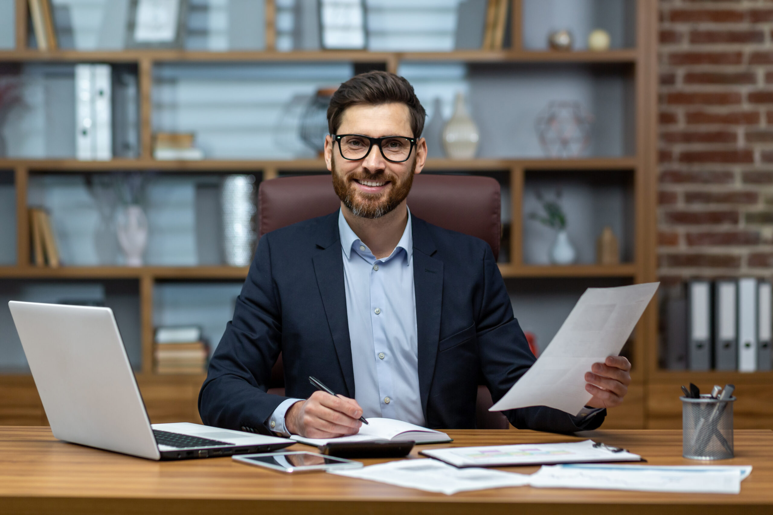 Portrait of a young handsome man, freelancer, businessman, accountant. Sitting in the office, looking at the camera, smiling. Works on a laptop, with documents, fills in forms with a pen